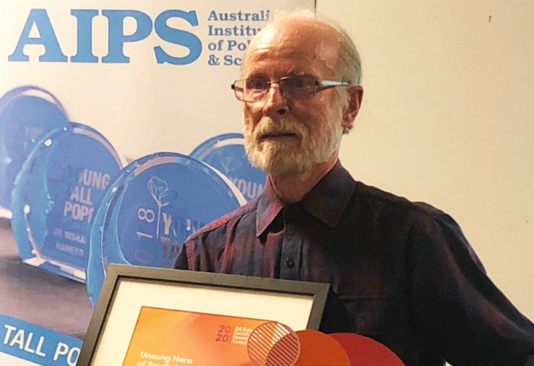 Mr Graham Medlin accepts his Unsung Hero of Science award. He holds a certificate in his arms and wears a dark shirt against a logo backdrop
